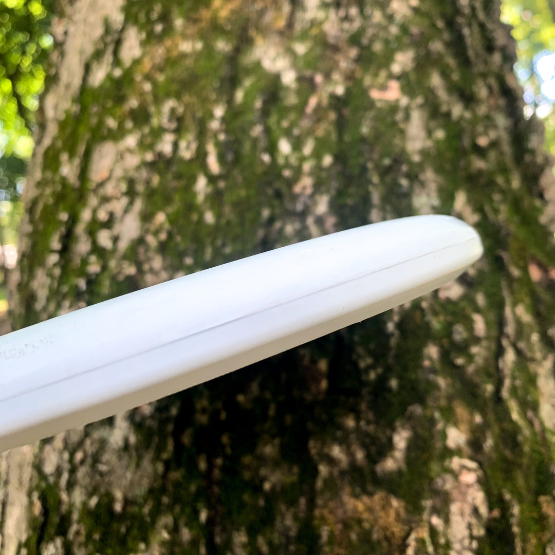 A white Magic - Suregrip - SSS putter disc clings to the trunk of a mossy tree, standing out against lush green foliage. This straight-flying putter is prized for easy control, even among vibrant surroundings.