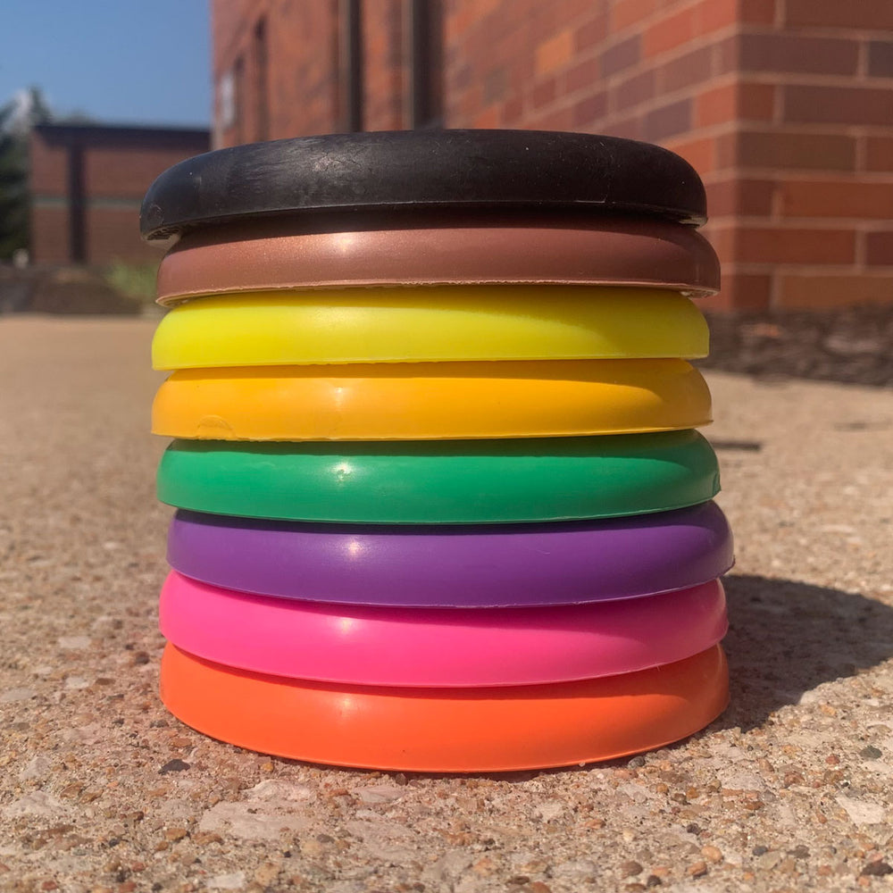 A stack of eight SureGrip™  Mini (Large) discs in assorted colors, displayed on a concrete surface near a brick building.