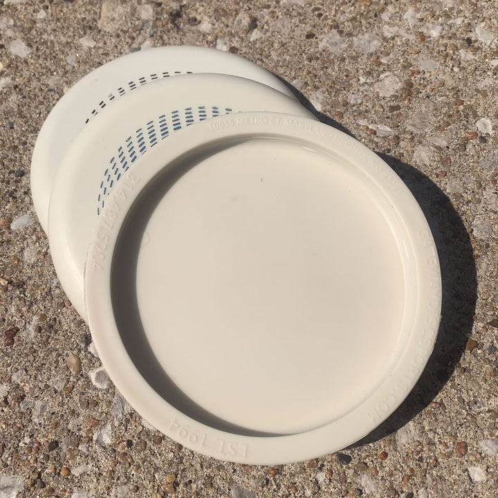 Three Mini (Large) - Superglow white plastic lids are stacked on a rough concrete surface in sunlight.