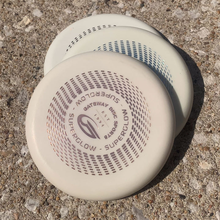 Two white Gateway Mini (Large) - Superglow discs are stacked on a rough concrete surface in sunlight.