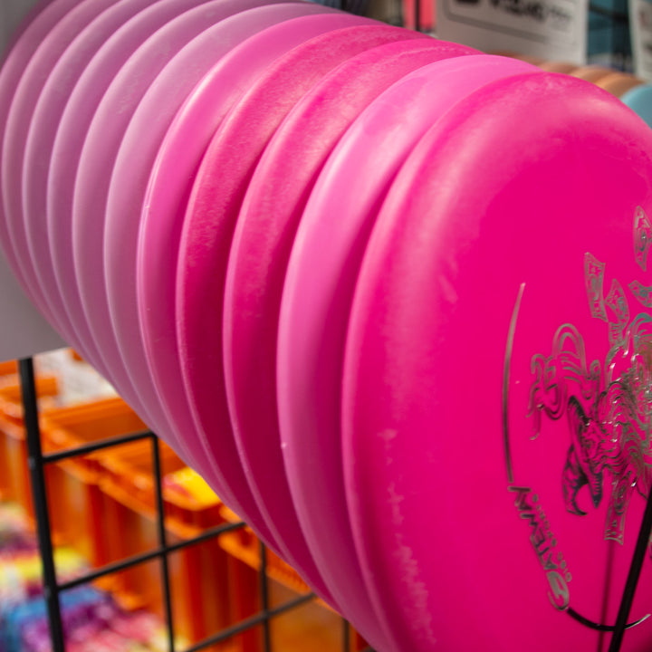 A close-up of several pink and purple Wizard - Money putters, a stable workhorse known for accuracy, stacked vertically in a metal display rack.