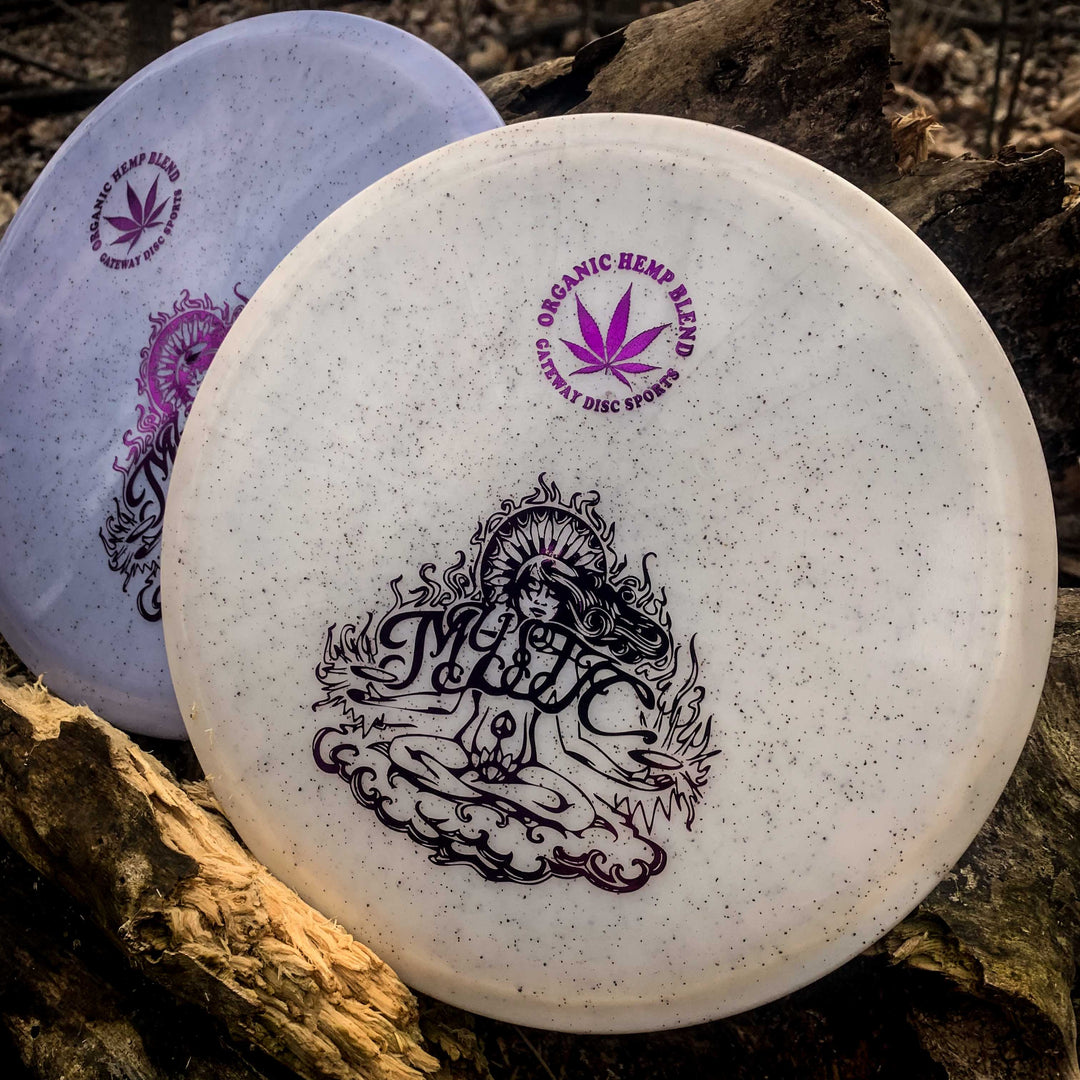 Two Mystic - Diamond Hemp disc golf discs rest on a log; both have speckled surfaces, detailed designs, and —Organic Hemp Blend—� text with a cannabis leaf, ideal for players wanting reliable straight mid-range throws.