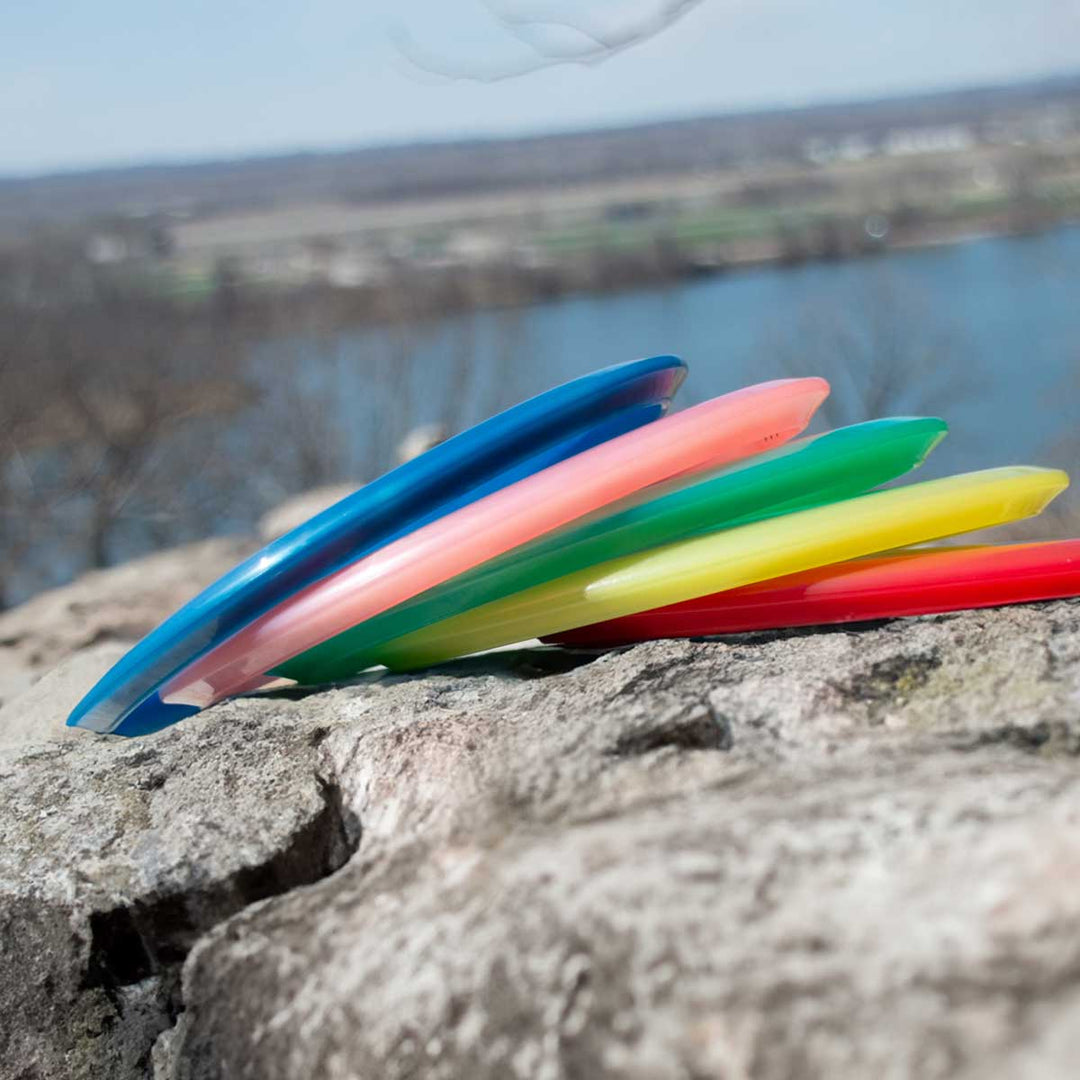 Five Element - Diamond mid-range discs in blue, pink, green, yellow, and red are stacked on rocks by a river with a scenic landscape behind them. Known for straight flight and excellent glide.