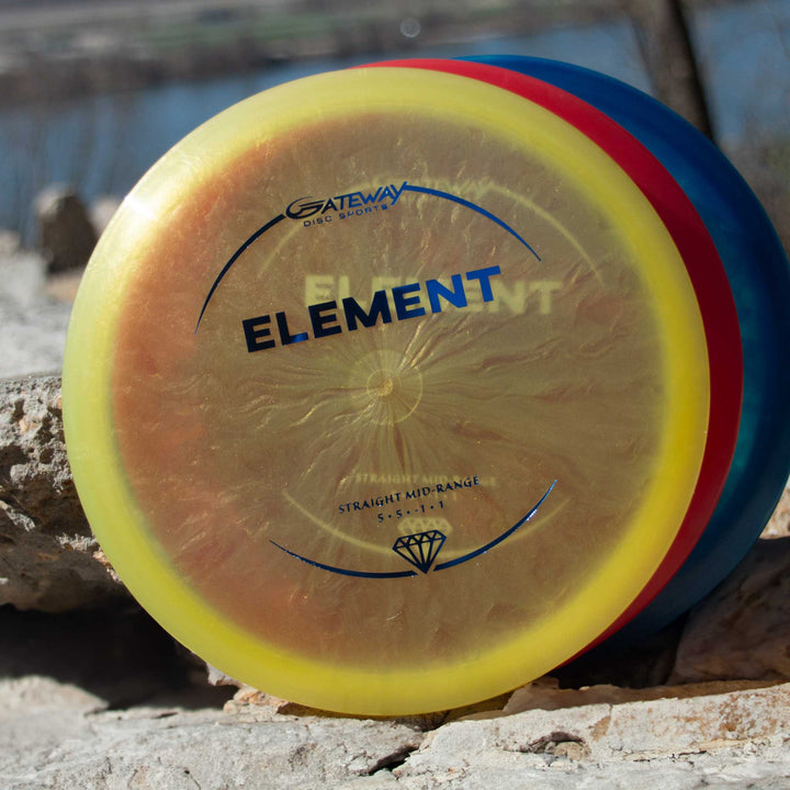 A Gateway Disc Sports Element - Diamond mid-range disc with a yellow and red rim and blue lettering, known for its straight flight, rests on rocks outdoors.