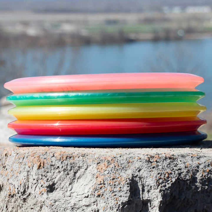 A stack of six vibrant Element - Diamond mid-range discs rests on rough concrete, offering straight flight and glide, with a blurred outdoor setting and water in the background.