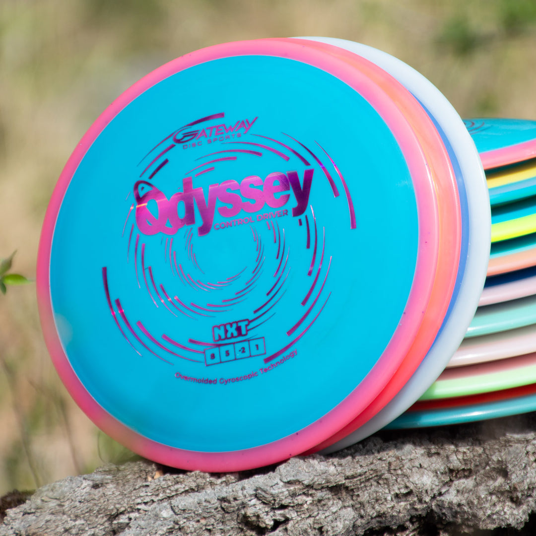 A stack of colorful disc golf discs sits on a tree trunk, topped by the Odyssey - NXT control driver, which features a blue, pink, and white design and advanced dual-material technology.