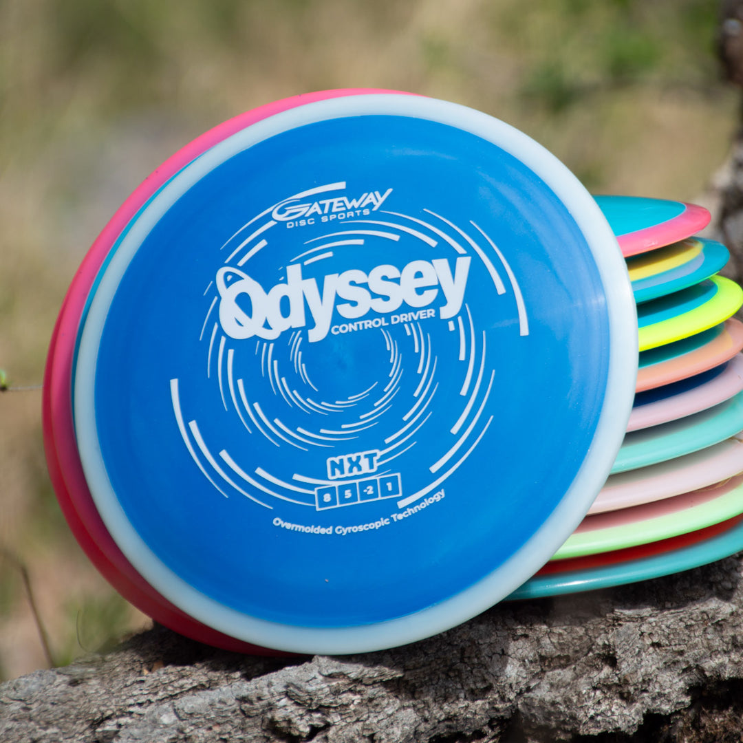 A stack of colorful disc golf discs is shown outdoors, with a blue Odyssey - NXT disc labeled "Control Driver" featuring dual-material technology prominently displayed on top.