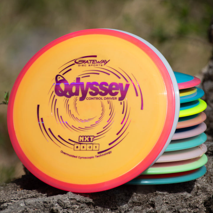 A stack of colorful disc golf discs, featuring the Odyssey - NXT control driver with dual-material technology by Gateway Disc Sports, rests on a tree stump outdoors.