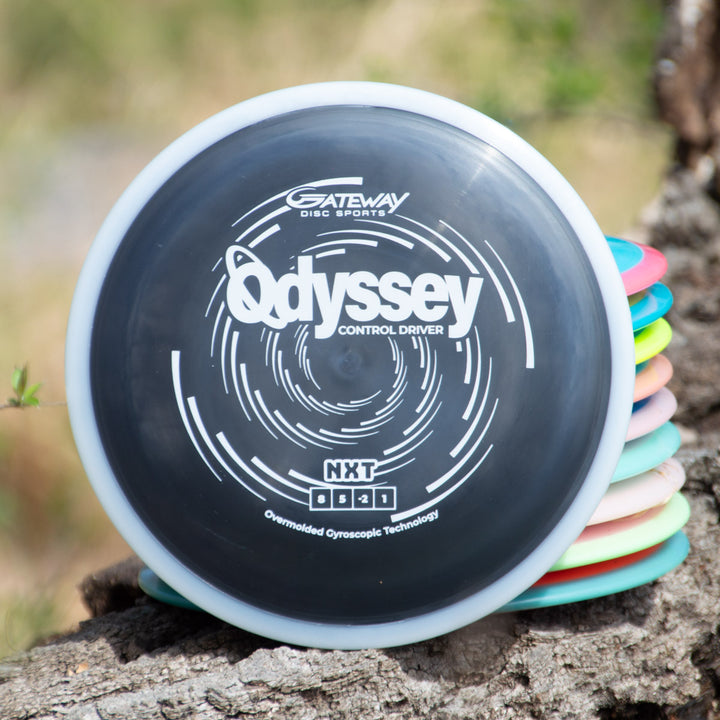 A stack of colorful disc golf discs is shown, topped by a black Odyssey - NXT Control Driver with dual-material technology from Gateway Disc Sports.