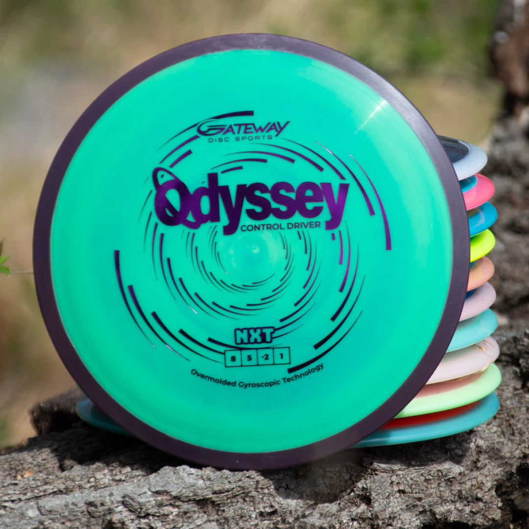 A stack of colorful disc golf discs sits on a tree trunk, topped by the green Odyssey - NXT disc control driver, which showcases advanced dual-material technology.