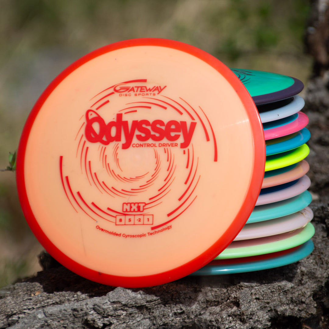 A stack of colorful disc golf discs sits on a tree stump, with the top disc—the Odyssey - NXT—featuring Gateway's innovative dual-material technology and labeled "Odyssey Control Driver.
