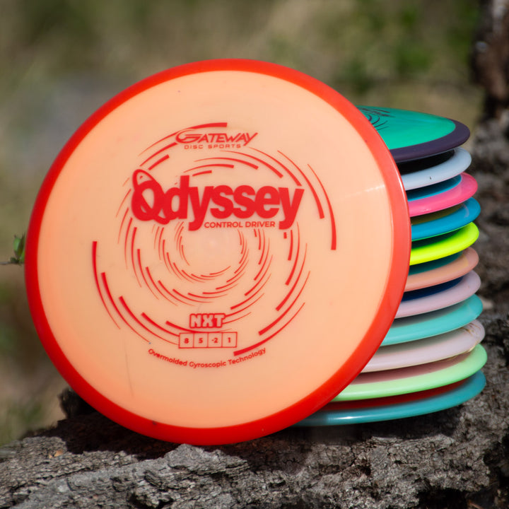 A stack of colorful disc golf discs sits on a tree stump, with the top disc—the Odyssey - NXT—featuring Gateway's innovative dual-material technology and labeled "Odyssey Control Driver.