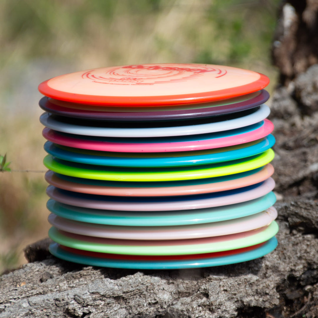 A stack of colorful Odyssey - NXT disc golf discs, featuring a control driver with dual-material technology, sits on a tree trunk outdoors, blurred grass and foliage in the background.