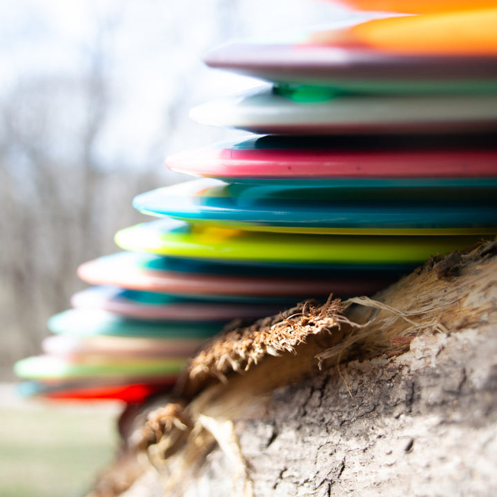 A stack of colorful Odyssey - NXT disc golf discs, featuring a control driver with dual-material technology, arranged on a rough tree trunk outdoors in a close-up shot with a blurred background.