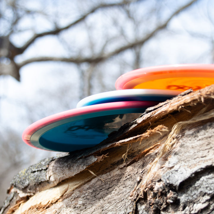 Three vibrant Odyssey - NXT discs, featuring control driver dual-material technology, rest on a broken tree trunk with blurred trees and sky in the background.