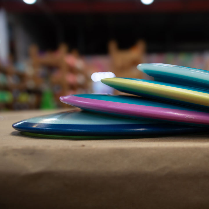 A stack of Odyssey - NXT discs with dual-material technology and blue, pink, and yellow edges rests on a brown indoor surface.