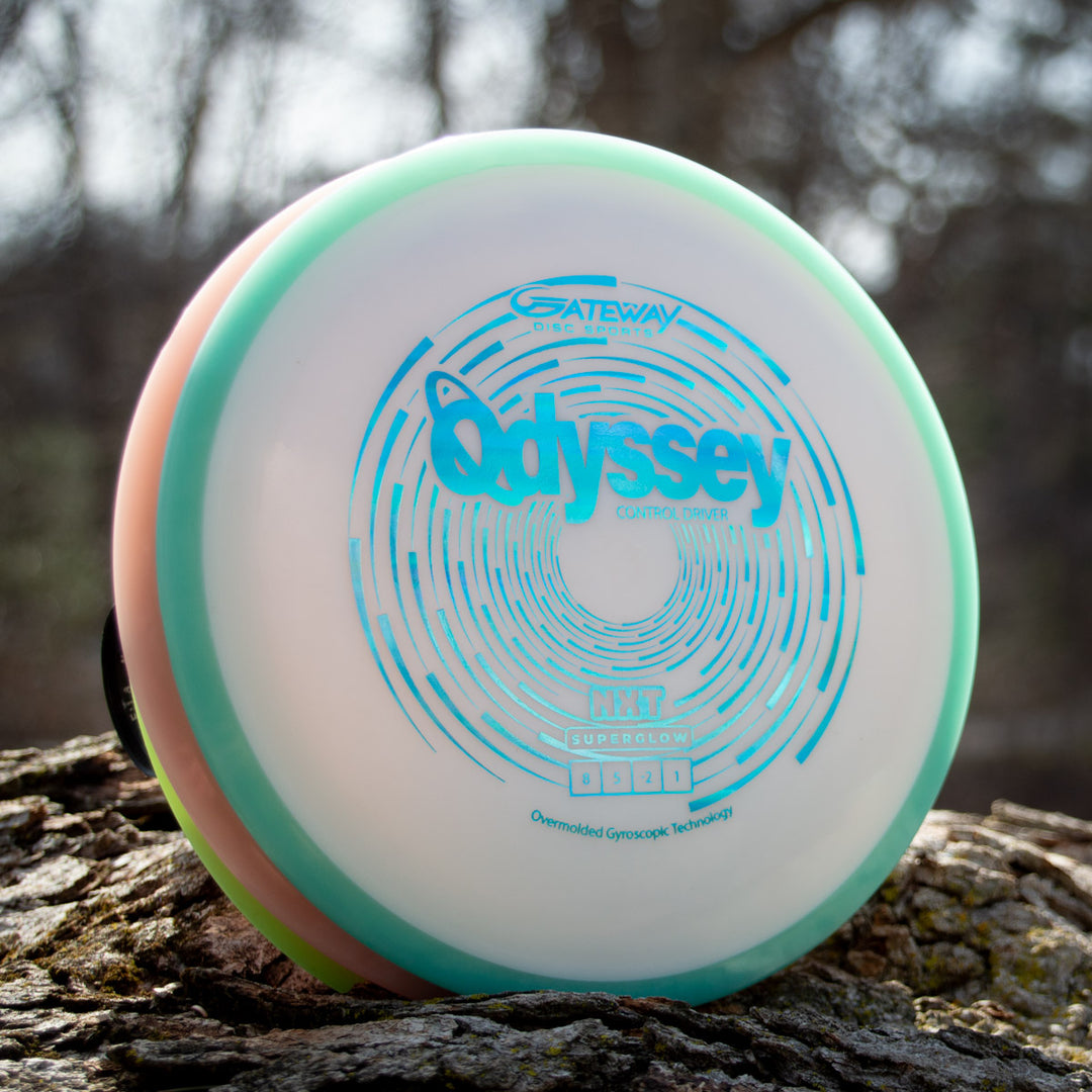 An Odyssey - NXT Superglow disc with teal and pink accents rests on a tree trunk outdoors, displaying its flight numbers and spiral design—bringing the spirit of David McCormack's Odyssey Story to every throw.