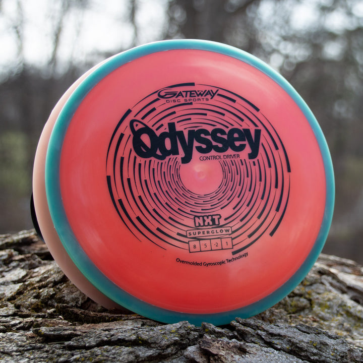 A pink and turquoise Odyssey - NXT Superglow disc golf driver stands upright on tree bark, displaying its flight numbers and other details—a striking nod to David McCormack's Odyssey Story.
