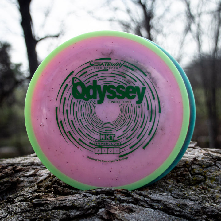 A pink and green disc golf disc labeled "Odyssey - NXT Superglow" rests on a tree trunk outdoors, echoing David McCormack's Odyssey Story, with softly blurred trees and sky in the background.