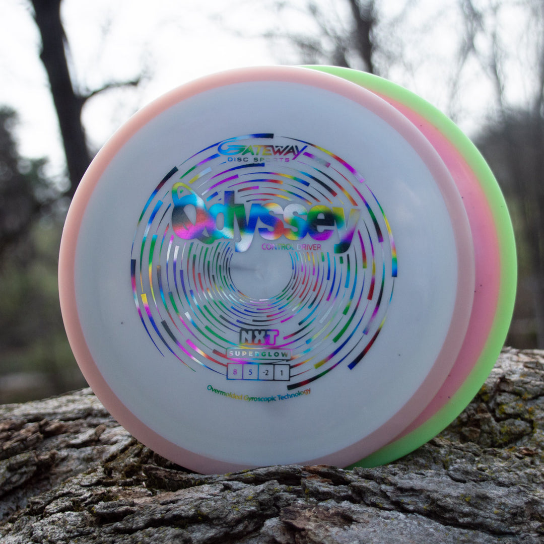Three colorful Odyssey - NXT Superglow disc golf discs with "Gateway Disc Sports" and "Odyssey" logos rest on a tree trunk outdoors as sunlight filters through the trees, evoking David McCormack's Odyssey Story.