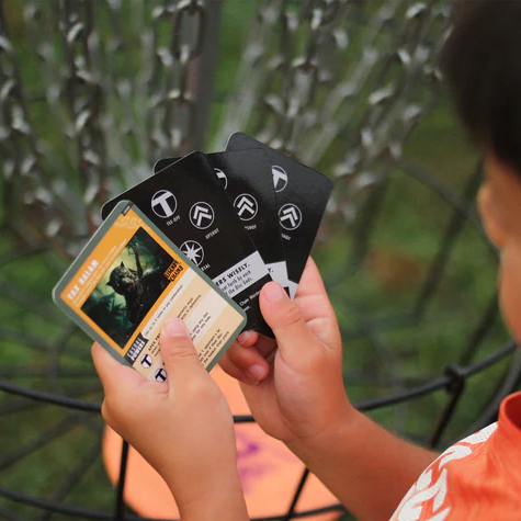 A child holds five playing cards—four black and one character card from Legends of the Chains: On-Course Disc Golf Game—while standing near outdoor disc golf chains.