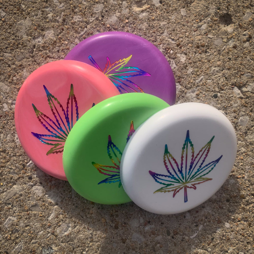 The Mini (Small) - SureGrip™ Leaf pins in purple, pink, green, and white, each featuring a holographic hemp leaf design, are shown displayed on a textured concrete surface.