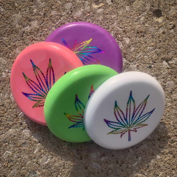 The Mini (Small) - SureGrip™ Leaf pins in purple, pink, green, and white, each featuring a holographic hemp leaf design, are shown displayed on a textured concrete surface.