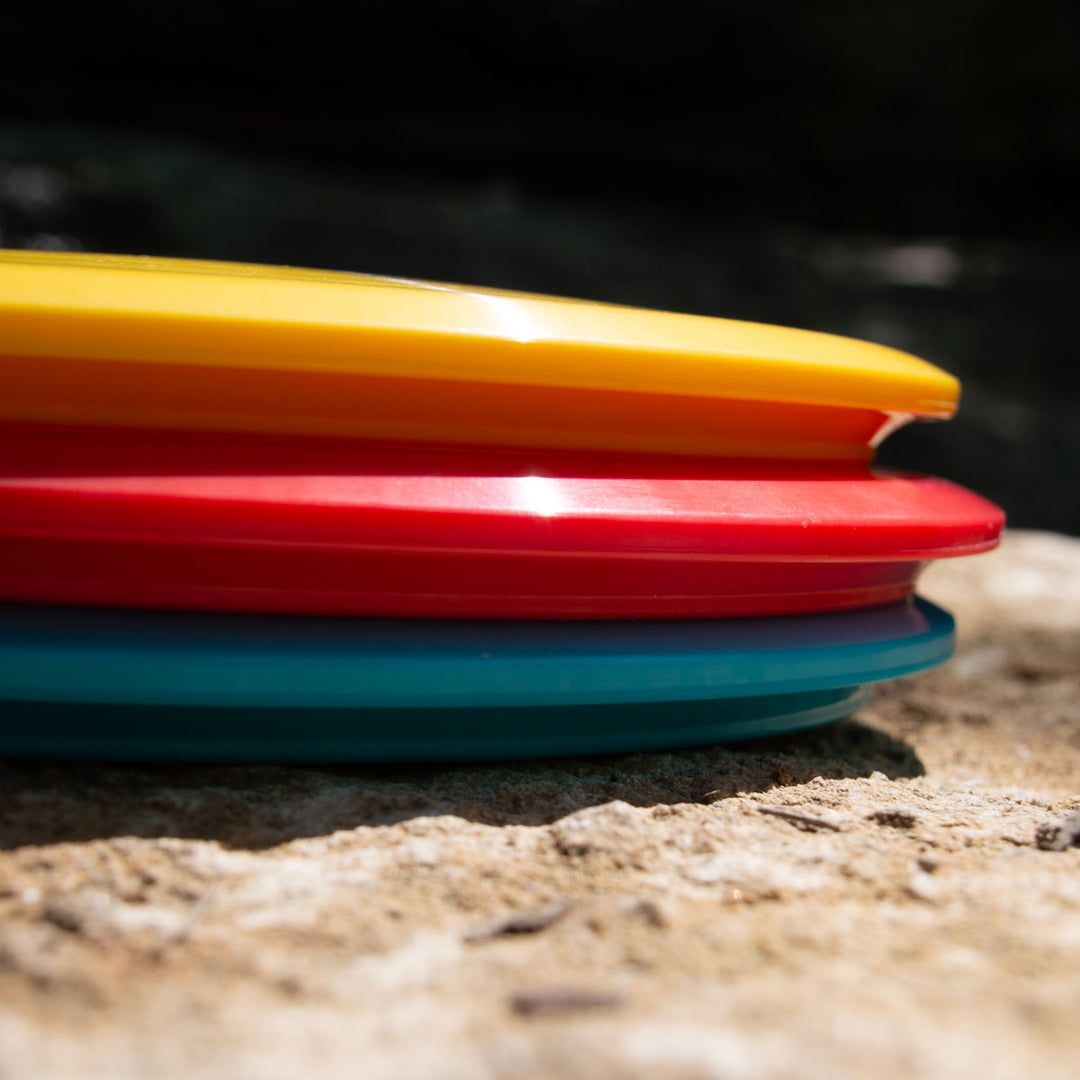 Close-up of stacked yellow, red, and blue Prophecy - NXT discs—crafted from NXT prototype material—resting on rough stone, set against a dark background.