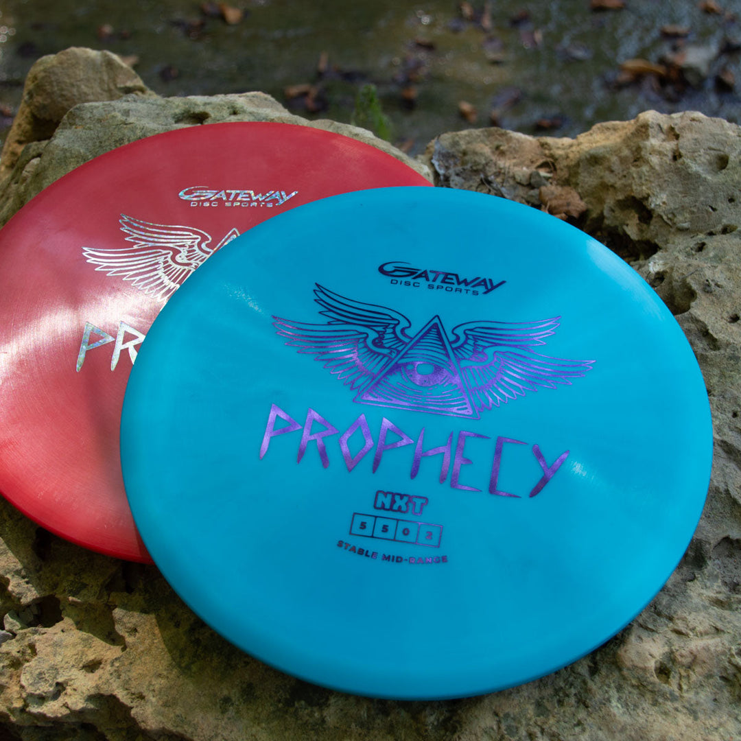 Two Prophecy - NXT discs—one red, one blue—sit on a large rock outdoors. Each overstable mid-range disc is made from NXT plastic and displays the unique eye-and-wings Prophecy logo.