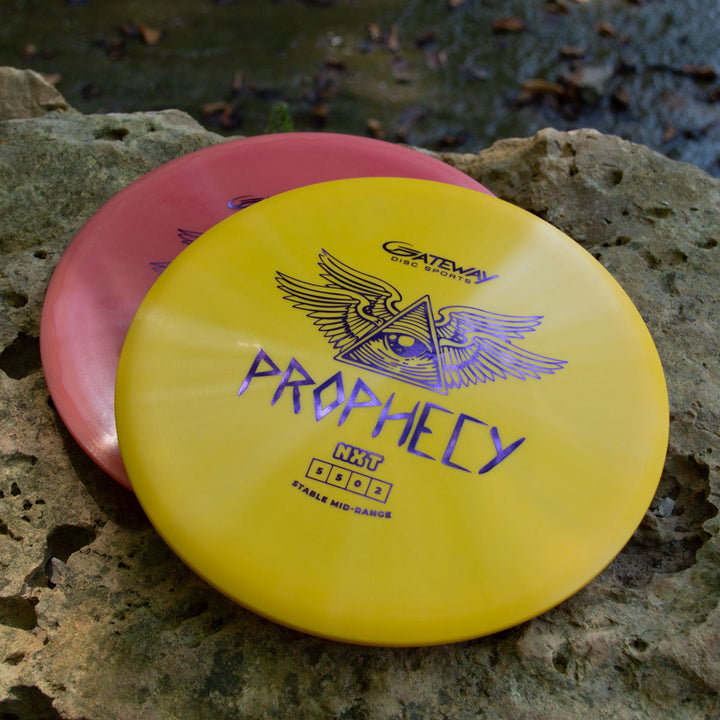 Two Prophecy - NXT discs rest on a rock outdoors: the top yellow NXT prototype displays an eye-in-triangle winged design, while the red disc below highlights this overstable mid-range's reliable flight on any throw.
