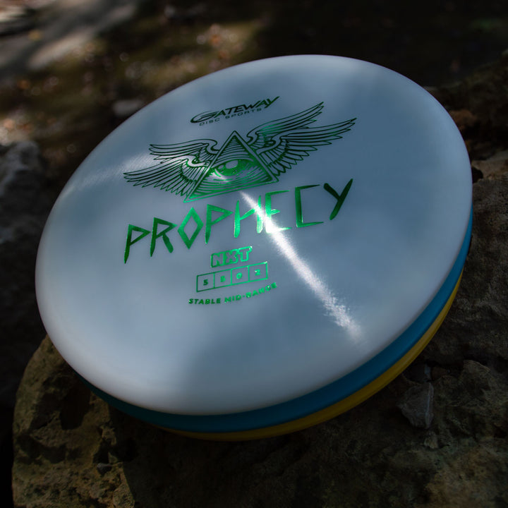 A white Prophecy - NXT overstable mid-range disc with green text and an eye-with-wings logo sits on a rock, crafted from prototype NXT material for consistent flight; other discs are partially visible beneath it.
