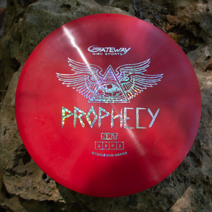 A red Prophecy - NXT disc by Gateway Disc Sports, made from durable NXT prototype plastic and displaying an eye-in-triangle-with-wings design, sits on rocks. This overstable mid-range ensures consistent flight on every throw.