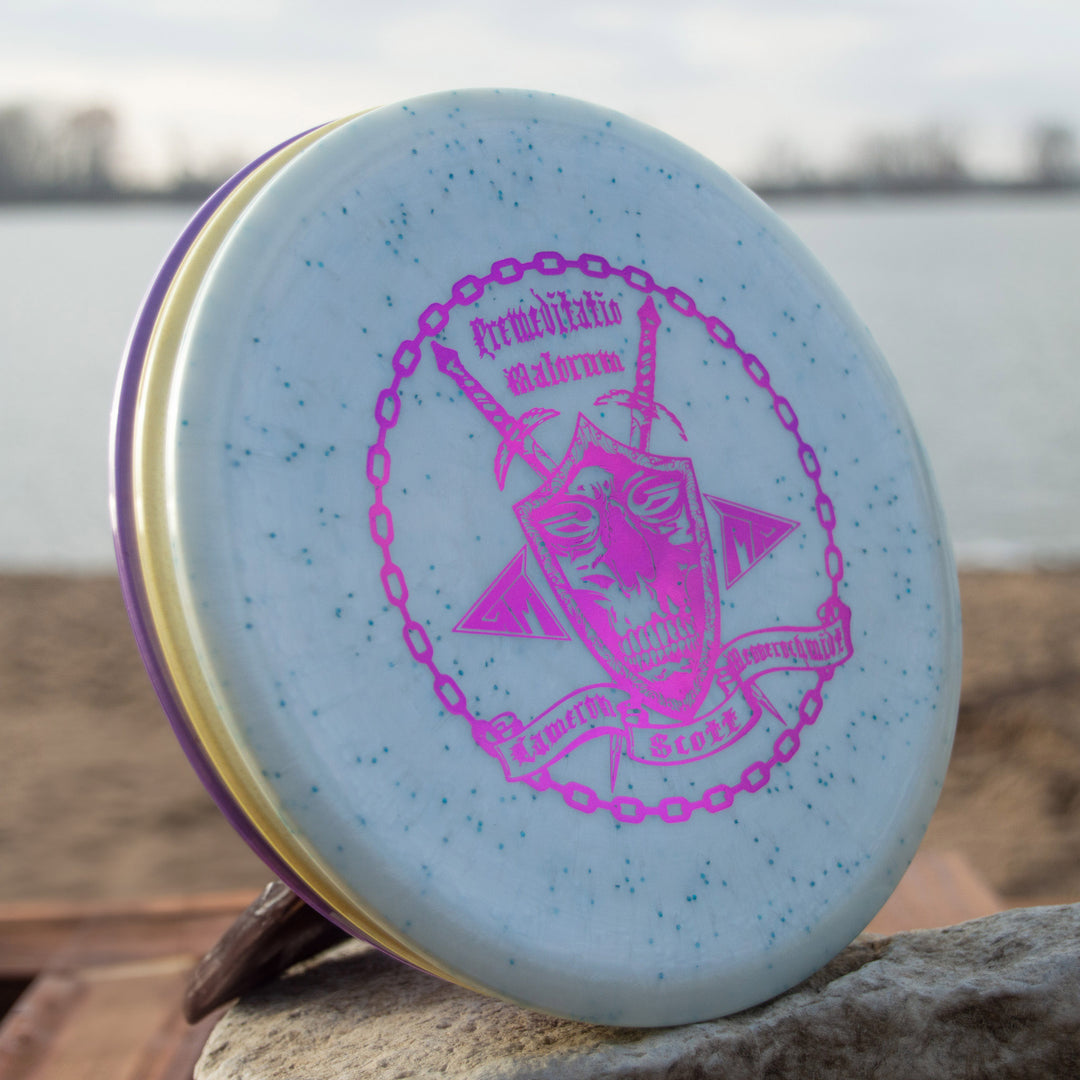 A light blue Cam's Prophecy - Diamond Metal Flake disc golf disc with purple speckles and a pink graphic lies on a rock near the sandy lakeshore. This overstable mid-range is ideal for controlled throws by the water.