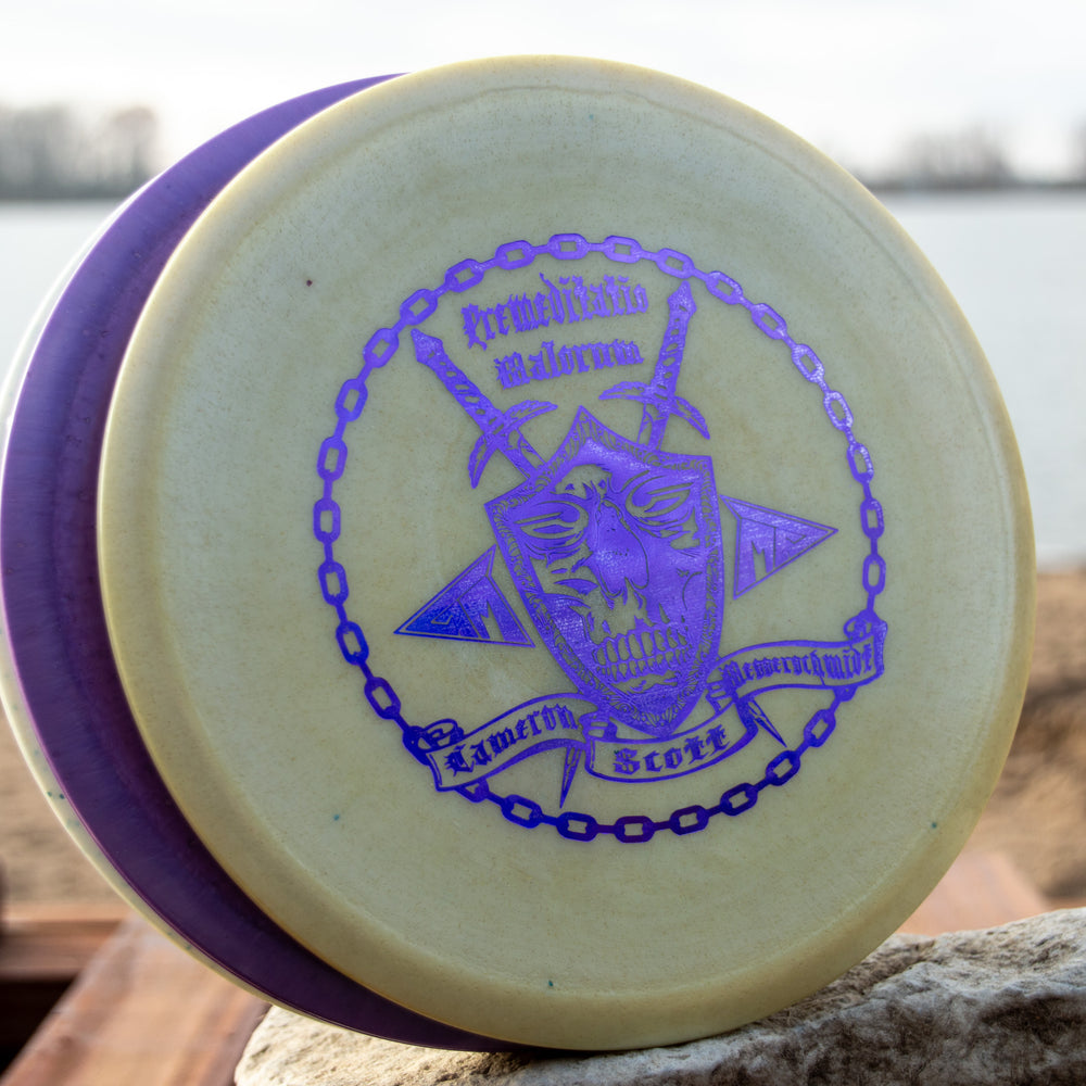 A Cam's Prophecy - Diamond Metal Flake disc, with a purple rim and metallic purple skull design, sits on a rock by the water—an eye-catching, overstable mid-range perfect for any disc golf collection.