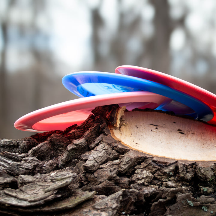 A vibrant Realm - Platinum distance driver disc golf disc sits on a freshly cut tree stump outdoors with blurred trees in the background, perfect for power throwers seeking reliable performance.