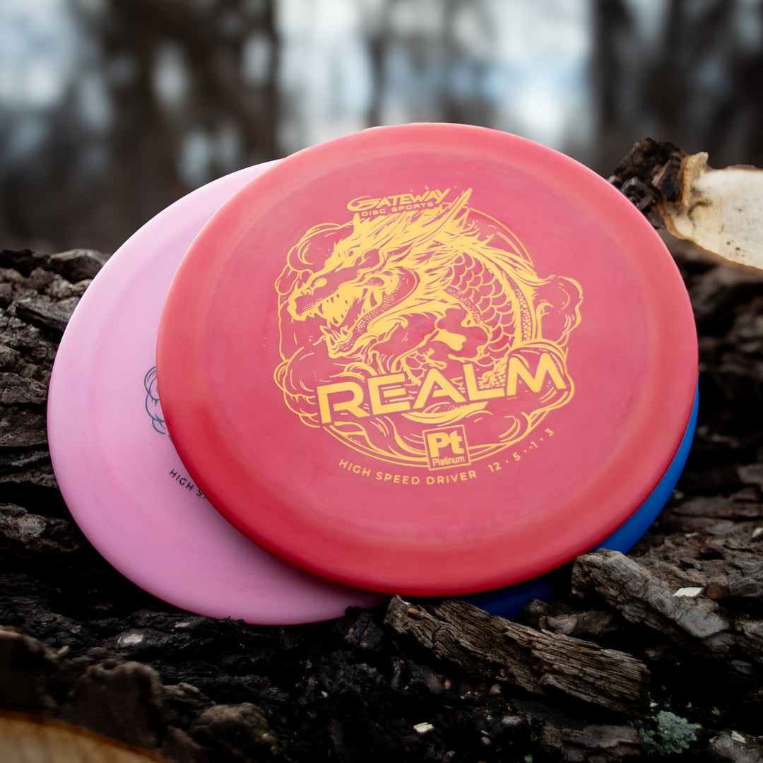Three disc golf discs rest on rough bark, the top one a Realm - Platinum distance driver with a gold dragon print—ideal for power throwers.