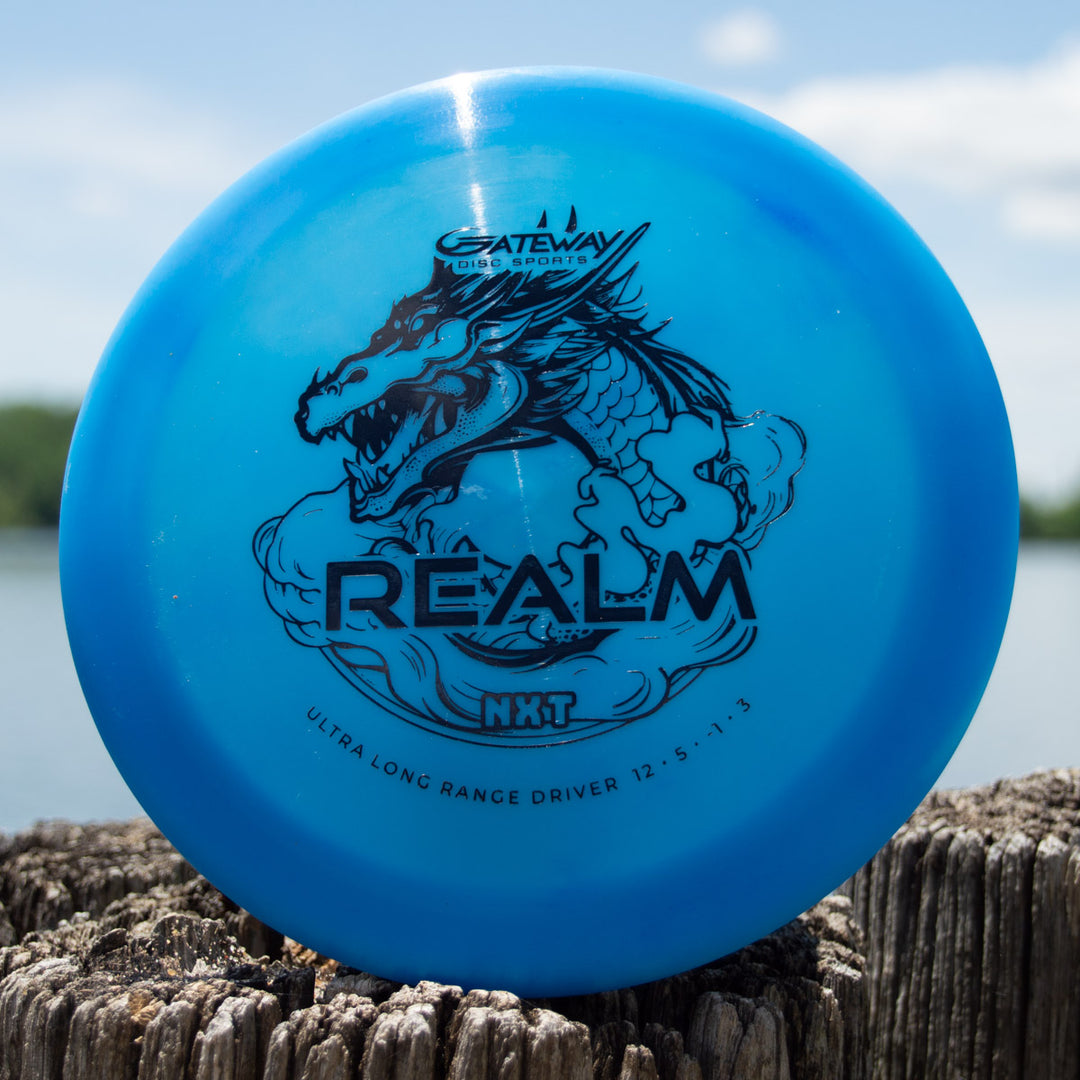 A blue "Realm NXT - Dragon" distance driver by Gateway Disc Sports rests on a weathered wooden post near water.