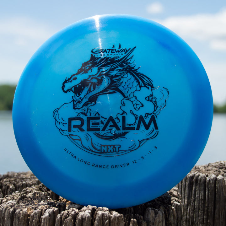 A blue "Realm NXT - Dragon" distance driver by Gateway Disc Sports rests on a weathered wooden post near water.