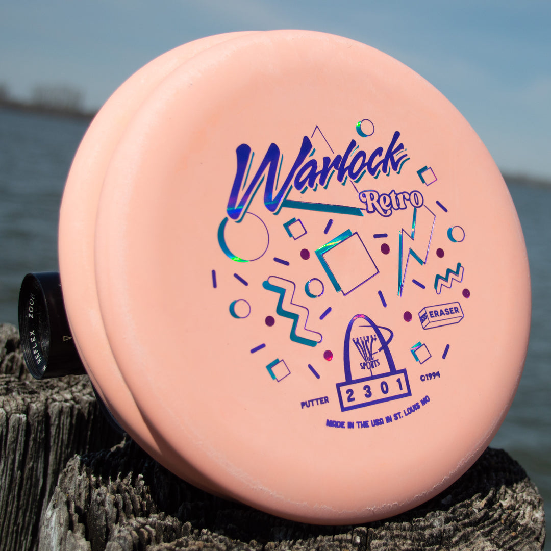 A Retro Warlock - Eraser disc golf putter in pink, featuring colorful geometric designs, sits on a weathered wooden post by the water.