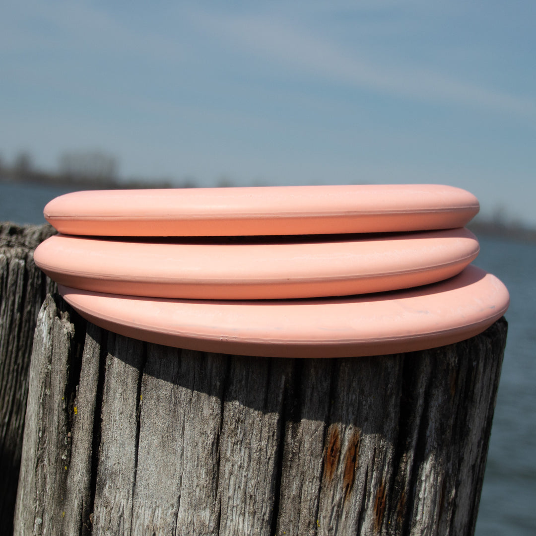 Three pink Retro Warlock - Eraser discs, including a putter, are stacked on a weathered wooden post near the water with a blurred shoreline in the background.
