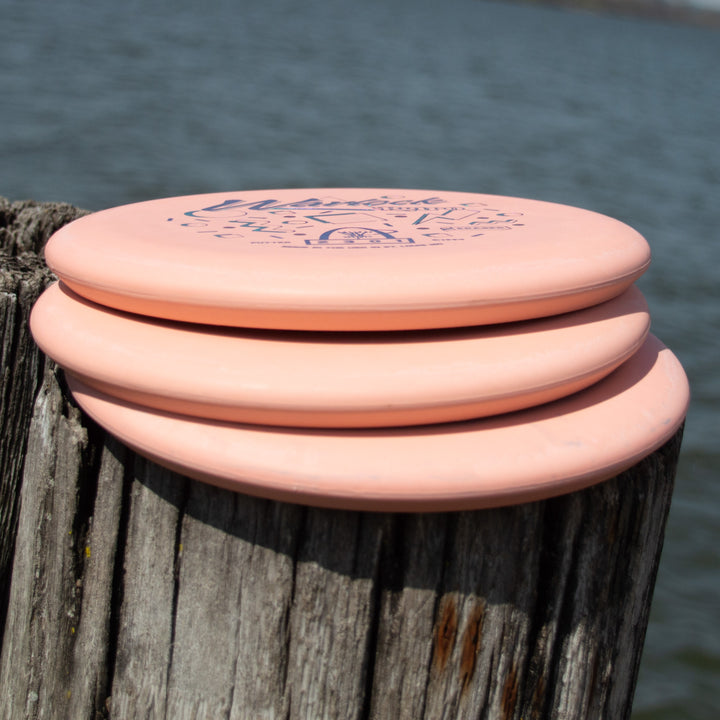 Three pink Retro Warlock - Eraser putters stacked on a weathered wooden post by the water.