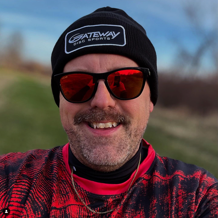 A person wearing a black Gateway Patch Beanie, red sunglasses, and a red patterned shirt smiles outdoors with a blurred natural background.