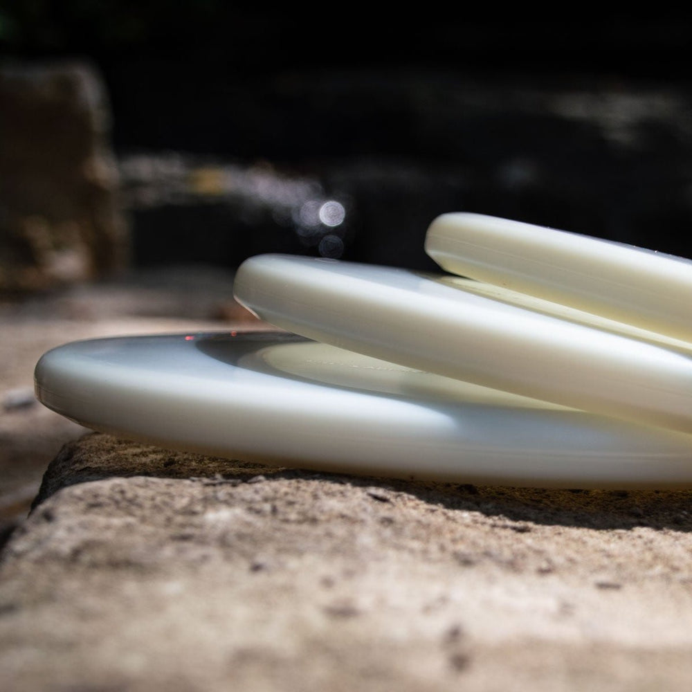 Three Shaman - Diamond Superglow disc golf discs, featuring a flat 25° nose and beardless rim, are stacked on stone outdoors, sunlight highlighting their edges with a blurred background.