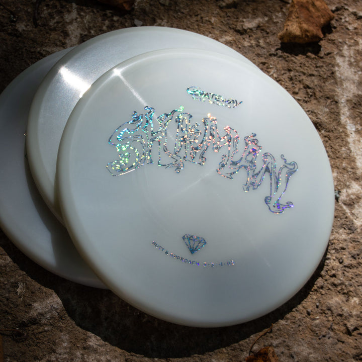 Three white Shaman - Diamond Superglow disc golf discs with iridescent Sky God IV text and Diamond Shaman logos sit on textured stone, their no-bead rim design illuminated by sunlight and surrounded by dried leaves.