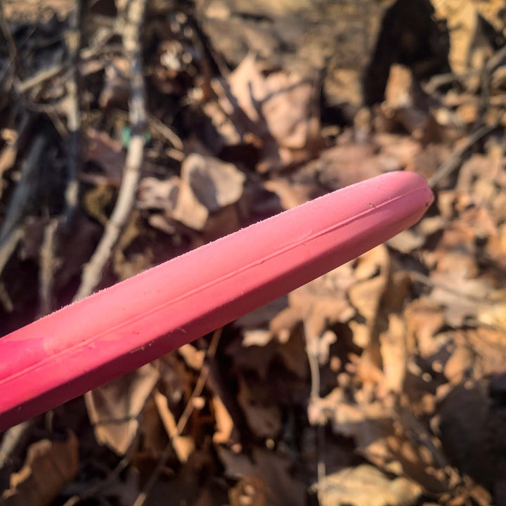 A close-up of a pink Shaman - Suregrip disc golf disc, featuring a beardless rim, held outdoors at an angle with dry leaves and sticks visible on the ground in the background.