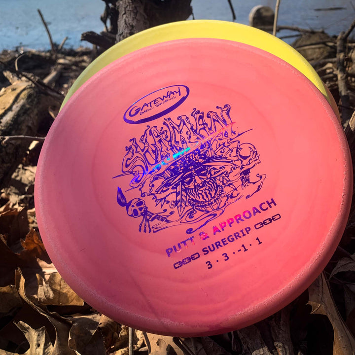 A close-up of a pink Shaman - Suregrip disc golf putter with graphics, featuring a flat 25-degree nose and beadless rim, resting on dry leaves near water.