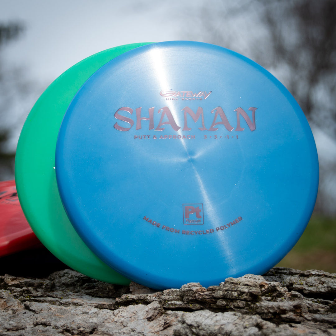 A blue Shaman - Platinum disc golf putter made from recycled plastic leans against a green and red disc, resting on a rock outdoors.