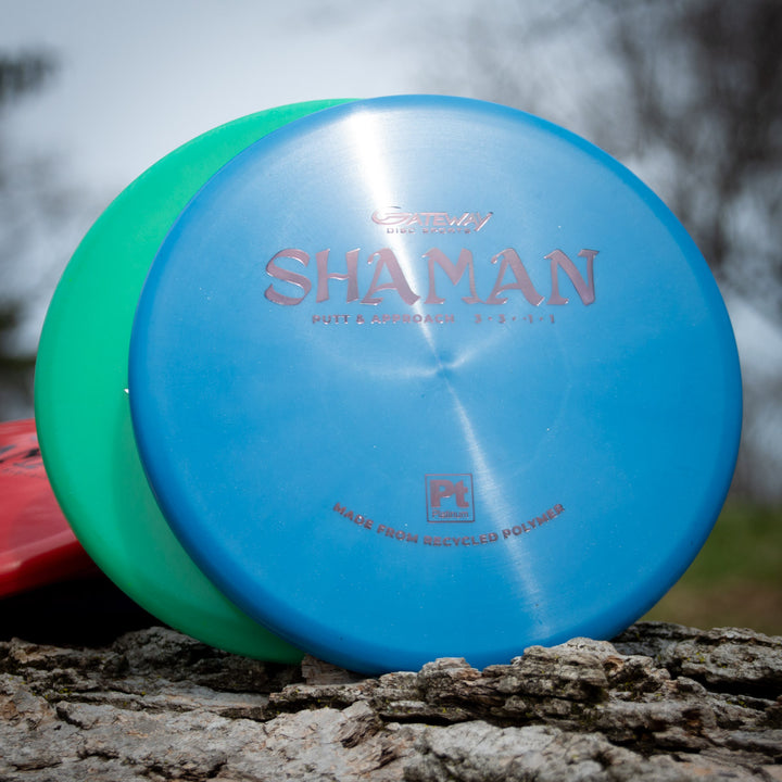 A blue Shaman - Platinum disc golf putter made from recycled plastic leans against a green and red disc, resting on a rock outdoors.