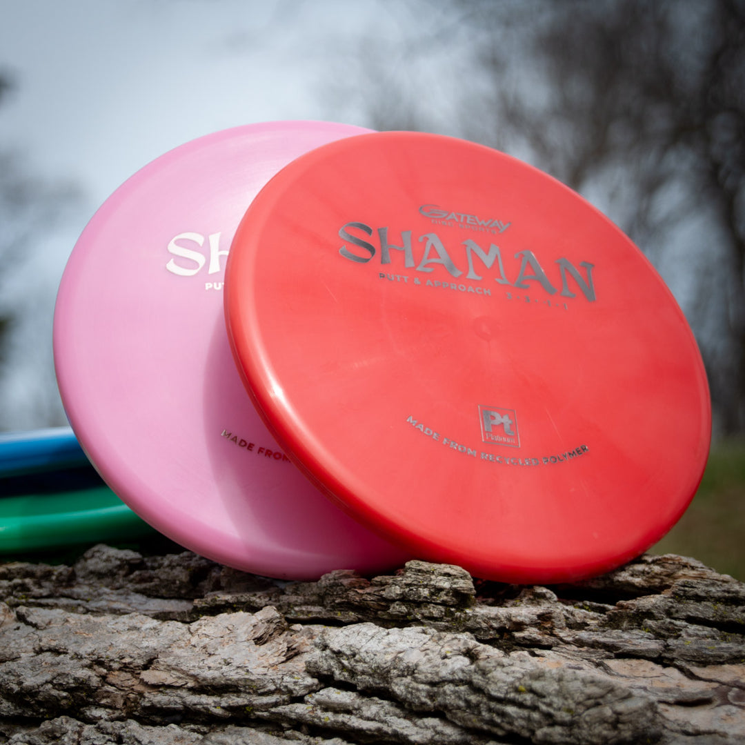 Two Shaman - Platinum discs in red and pink recycled plastic stand upright on a rough tree trunk outdoors, with blurred trees behind—a sustainable choice for eco-conscious disc golf players.
