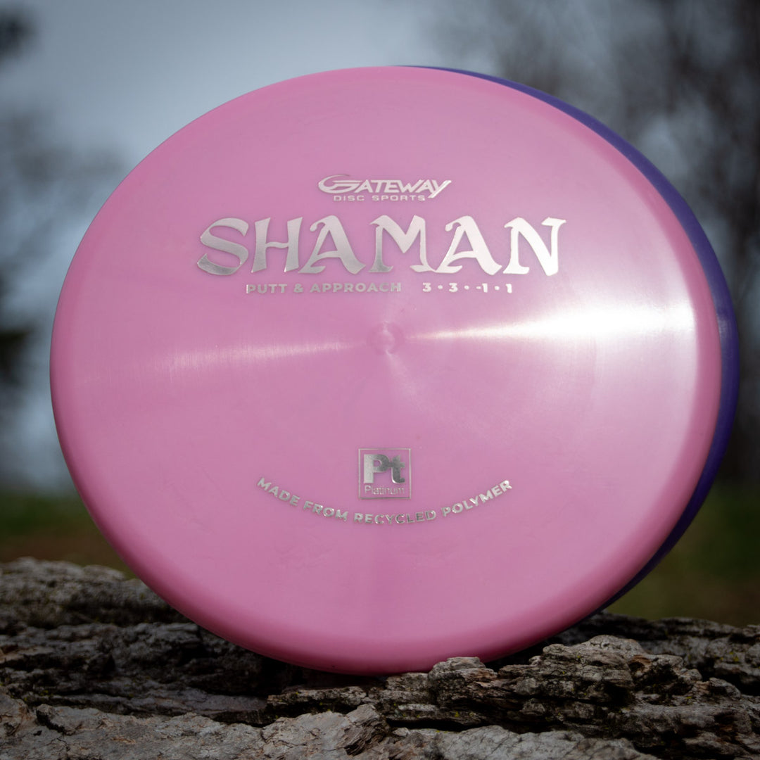 A pink Shaman - Platinum disc golf putter made from recycled plastic rests on a tree stump, its silver text highlighting its sustainable design.